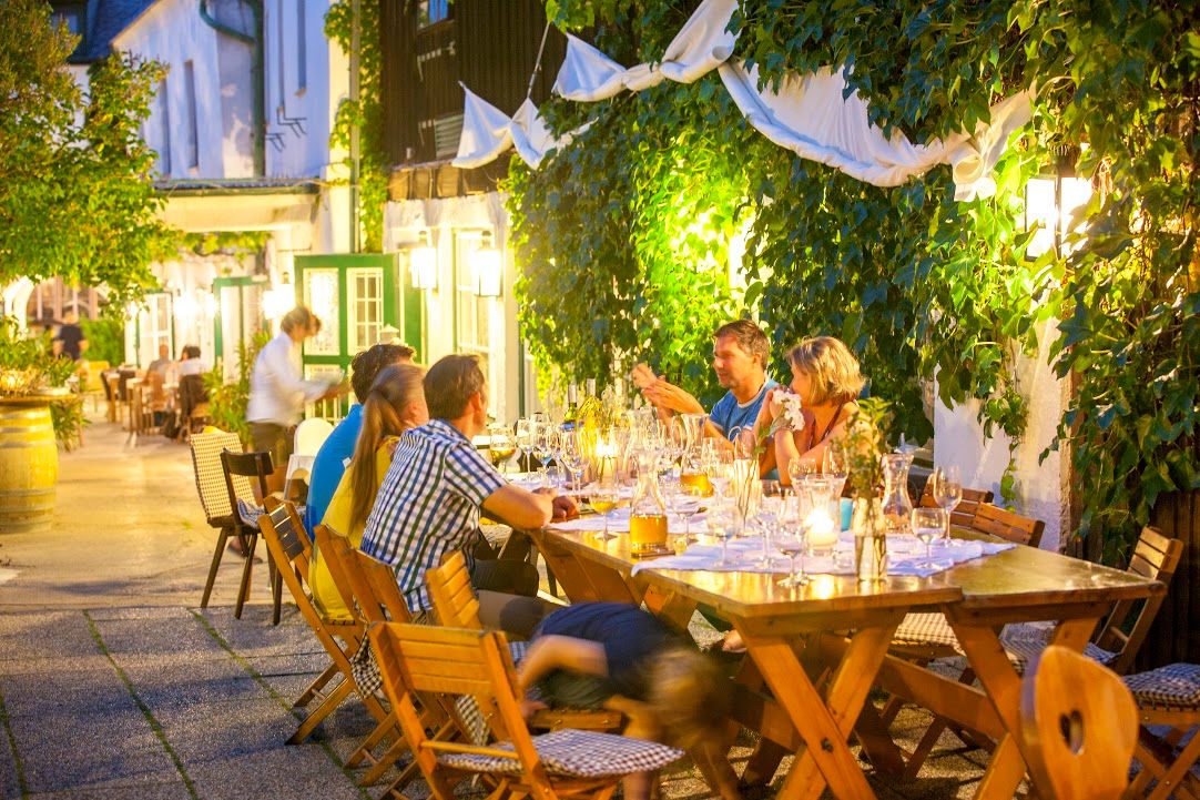 Menschen sitzen an einem langen Tisch im Freien, umgeben von Pflanzen und Lichtern.